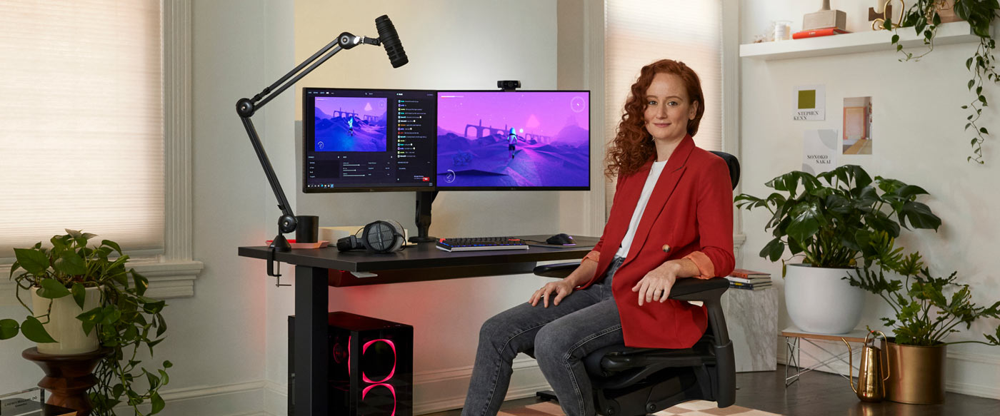 Foto de la Dra. Lindsey Migliore sentada en una silla gaming Embody frente a la cámara con un escritorio gaming Nevi en negro con brazos para monitor Ollin