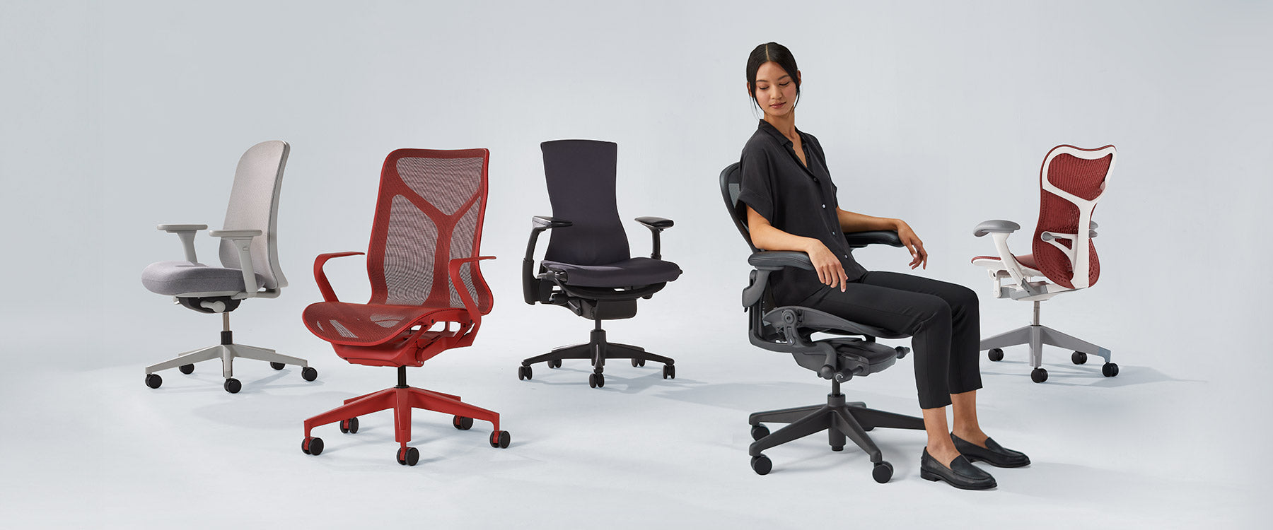 Foto de sillas Herman Miller alineadas, entre las que se incluyen Verus en blanco mineral y gris, Cosm con respaldo medio en rojo cañón, Embody en negro, Aeron en grafito con una mujer sentada y Mirra 2 en rojo cordobés
