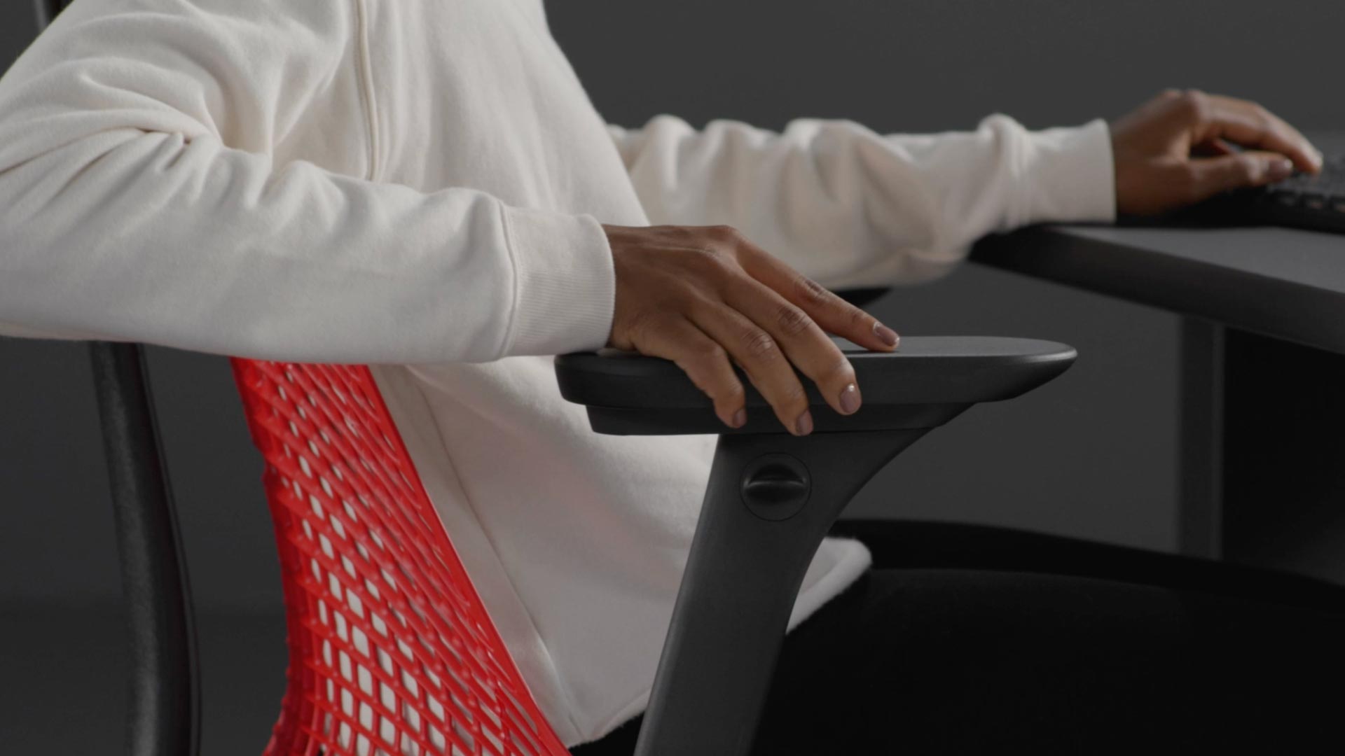 Vídeo de una persona sentada en una silla de juego Sayl roja y negra de Herman Miller, estirando la mano junto a la silla para ajustar los brazos.