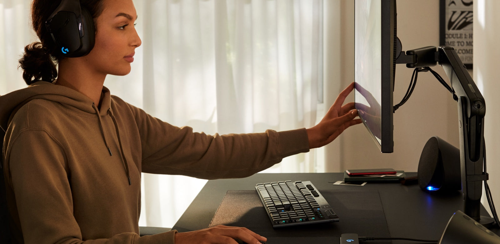 Una mujer sentada en un escritorio de juego Nevi negro con un brazo de monitor de juego Ollin negro de Herman Miller. Lleva auriculares Logitech y mueve la pantalla del ordenador con una de sus manos.