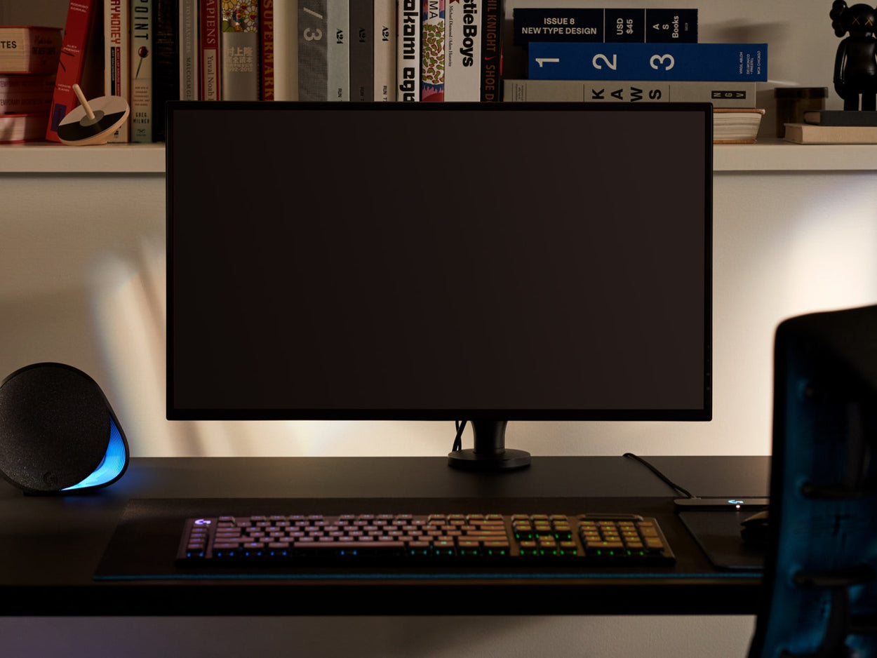 Vista frontal de un brazo de monitor Ollin negro que sujeta una pantalla de ordenador y se acopla a un escritorio de juego Nevi negro de Herman Miller. Ambientado en una sala de juegos o un despacho en casa, con una estantería, un teclado y un ratón.
