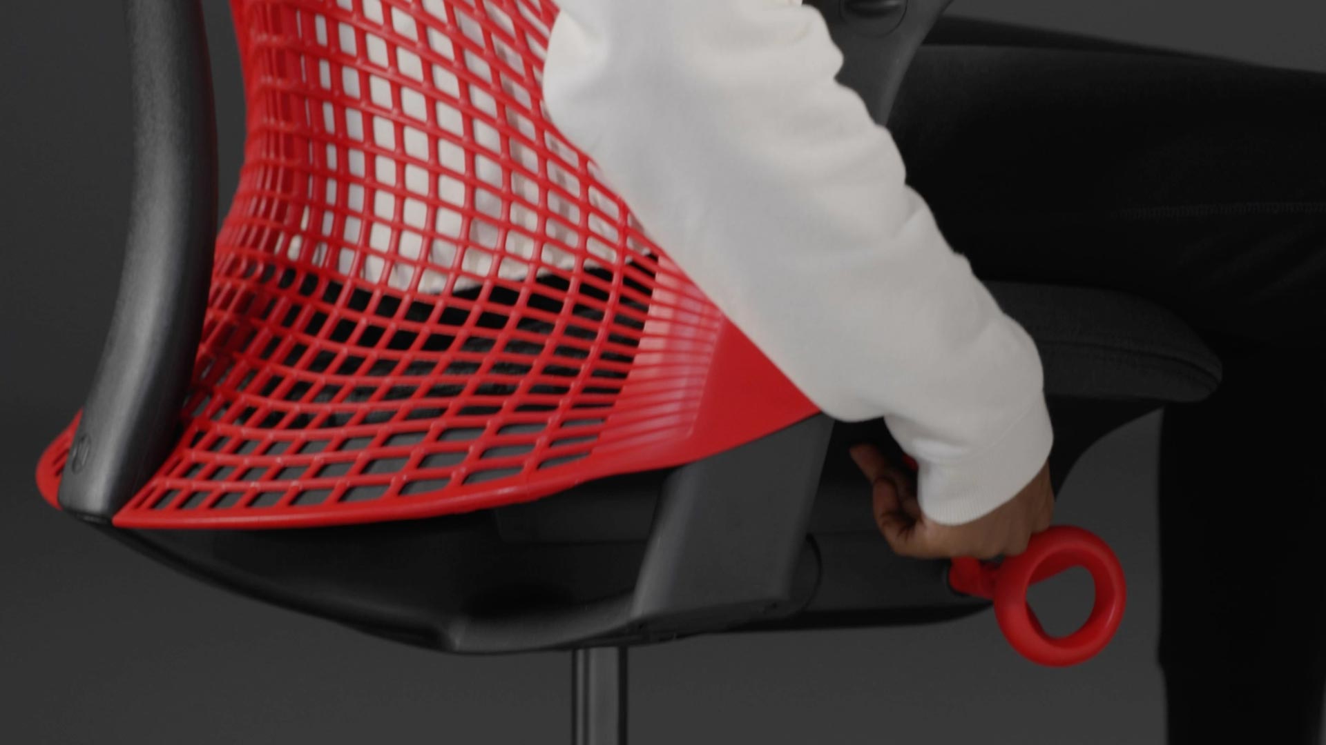 Vídeo de una persona sentada en una silla de juego Sayl roja y negra de Herman Miller, acercándose a la silla para ajustar la altura del asiento.