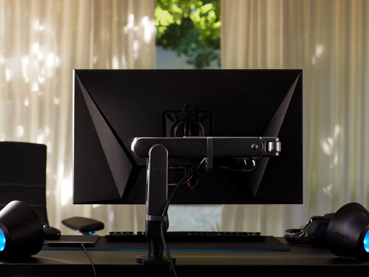 Vista trasera de un brazo de monitor Ollin negro que sostiene una pantalla de ordenador y está unido a un escritorio de juego Nevi negro de Herman Miller. Conjunto en una sala de juegos u oficina en casa, con luces, teclado y ratón.