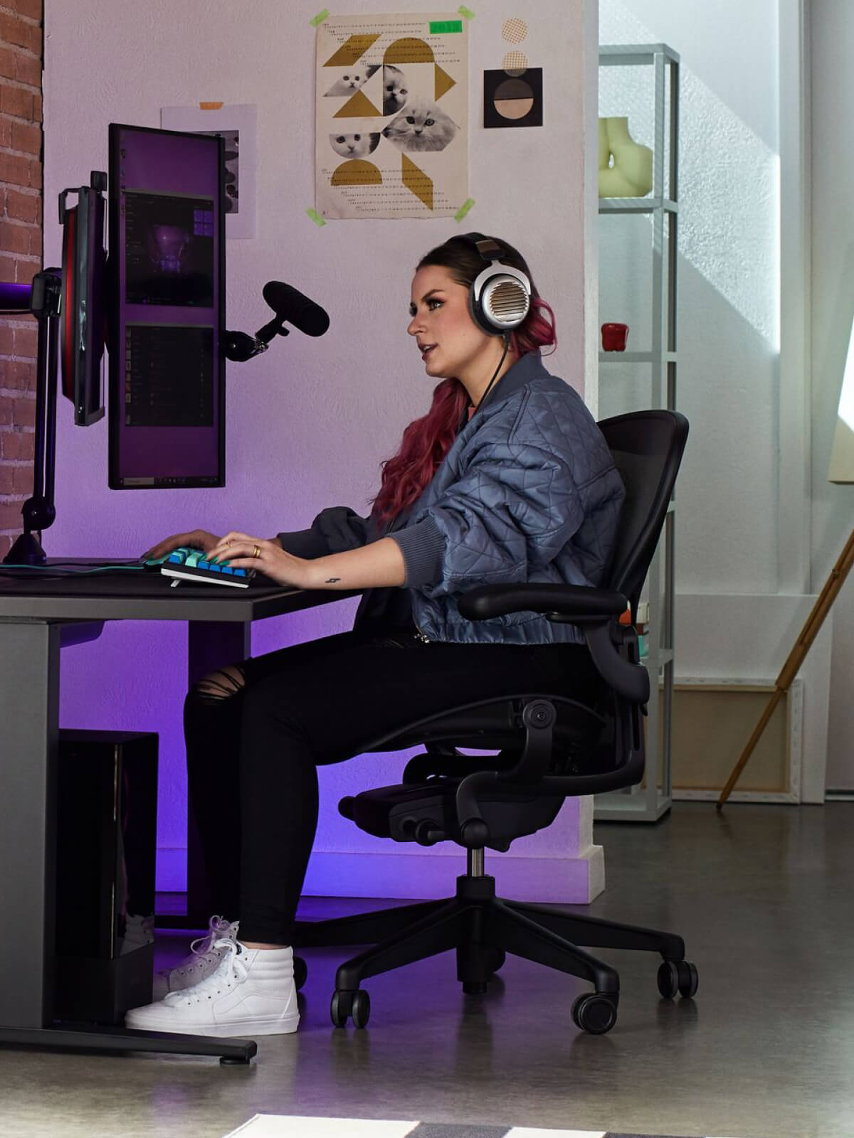 Electra jugando sentada en una silla Aeron en negro ónix con un escritorio para juegos Nevi en negro y un monitor Ollin