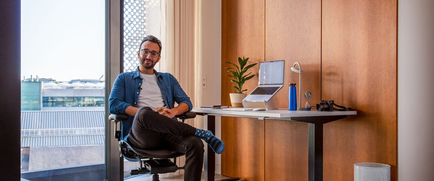 Foto de Ali Abdaal, podcaster, sentado en una silla de oficina Herman Miller Aeron en grafito frente a un escritorio sit-stand Nevi con patas negras y tablero blanco con un ordenador portátil en un soporte para portátiles Oripura