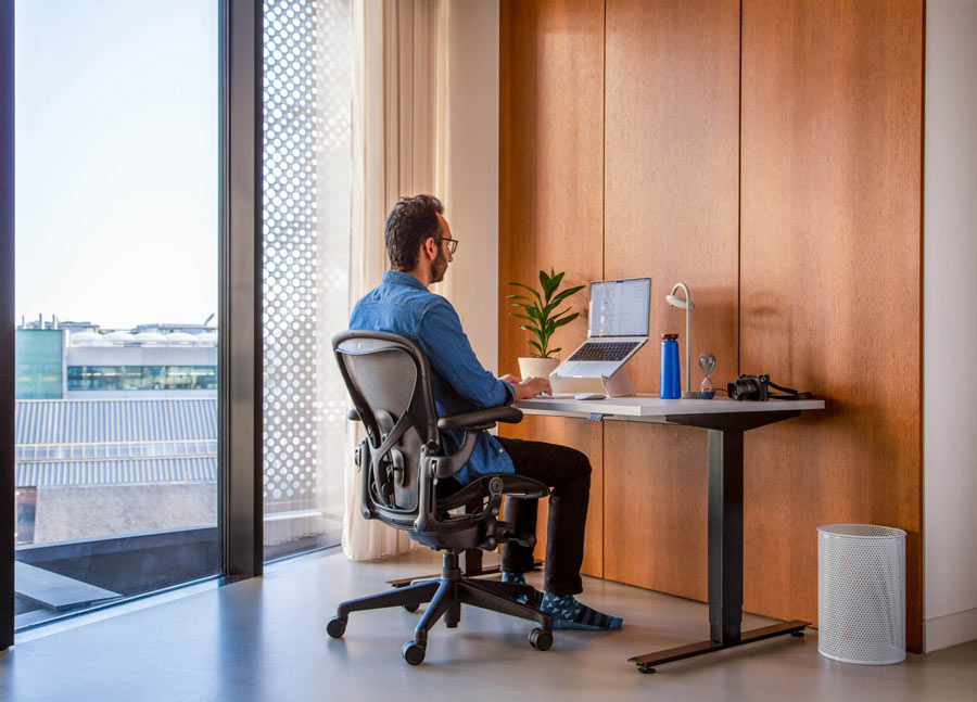 Foto de Ali Abdaal, podcaster, sentado en una silla de oficina Herman Miller Aeron en color grafito frente a un escritorio Nevi con patas negras y superficie blanca, con un ordenador portátil en un soporte Oripura.
