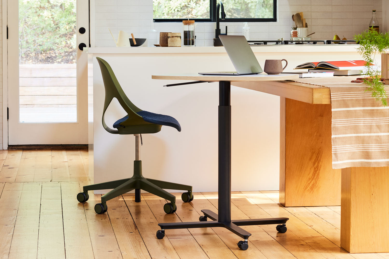 Foto tomada en una cocina doméstica con silla de oficina Zeph en color oliva con cojín de asiento en azul medianoche y mesa de trabajo compacta Passport de altura ajustable