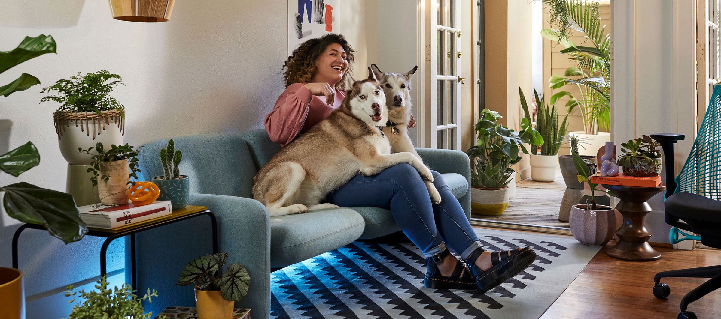 Friskk sentada en el sofá Wilkes en verde con sus dos perros, la silla gaming Sayl en negro y azul océano a un lado