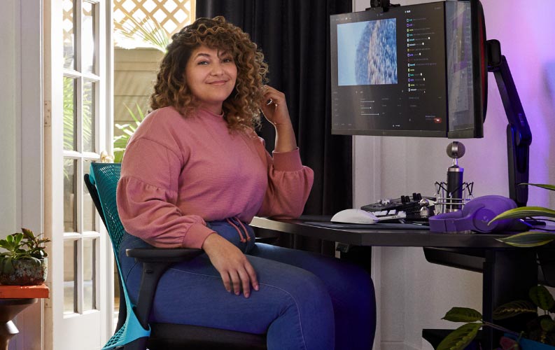 El streamer profesional y actor de doblaje Friskk sentado en una silla gaming Herman Miller Sayl en azul océano y negro, en una mesa gaming sit-stand Nevi negra con monitor en brazo para monitor Oliin.