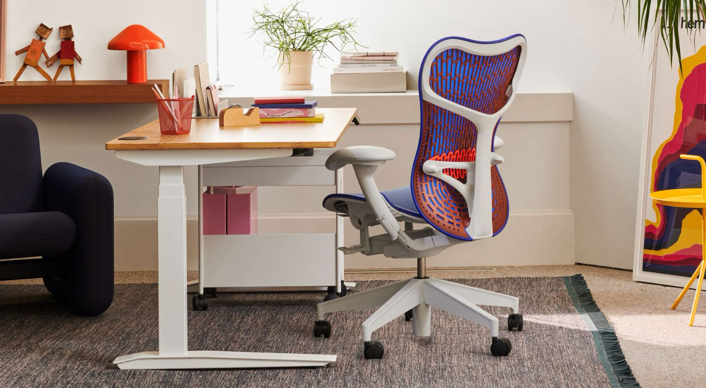 Una silla de oficina Mirra 2 azul, naranja y blanca de Herman Miller, colocada junto a un escritorio de pie Jarvis. Entorno de oficina en casa con un sofá Wilkes azul, una alfombra Maharam azul, una mesa auxiliar DLM amarilla y una lámpara de mesa HAY Pao roja.
