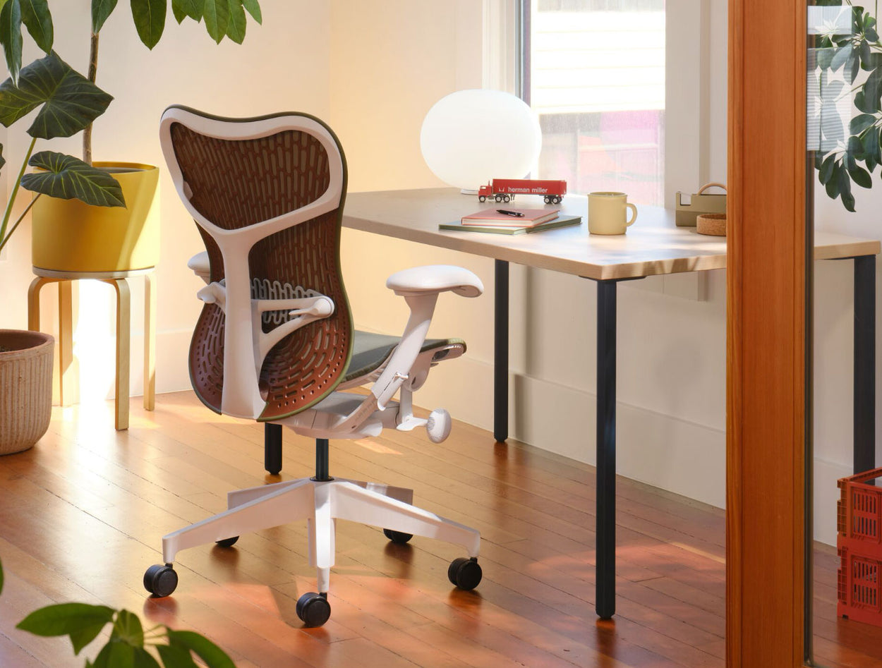 Una silla de oficina Mirra 2 verde oliva y blanca con respaldo granate, colocada frente a un escritorio OE1 de Herman Miller. En un entorno de oficina doméstica, con suelo de madera maciza, paredes blancas y una iluminación cálida y acogedora.