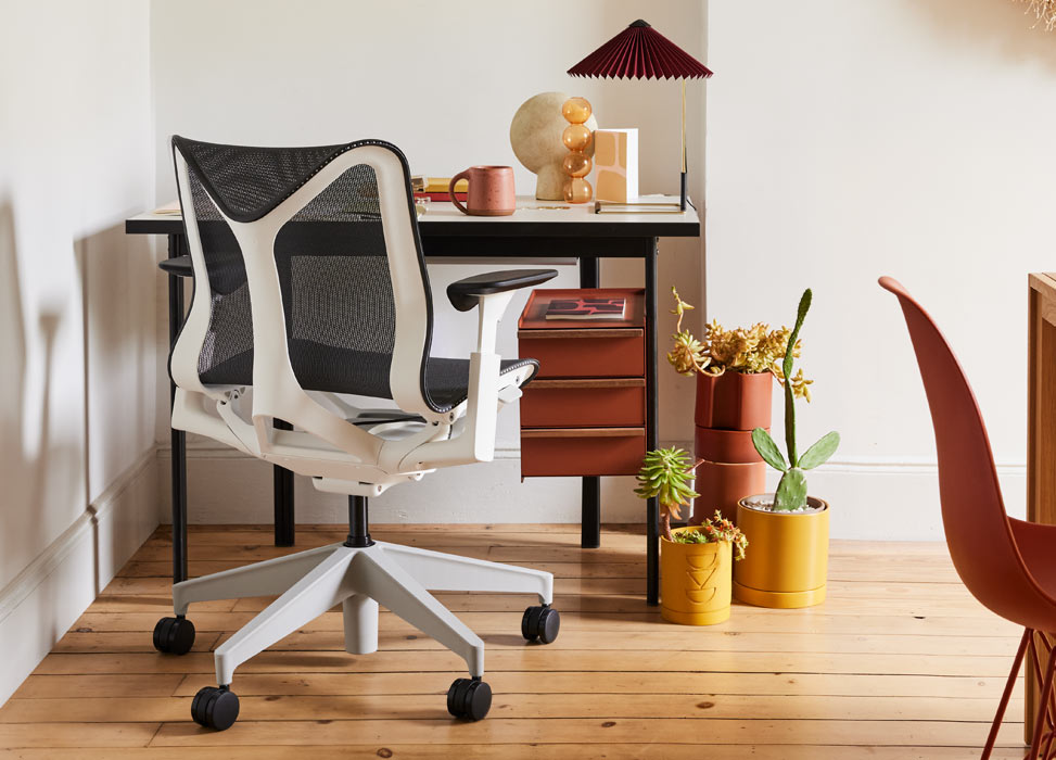Una silla de oficina Cosm azul y blanca de Herman Miller, colocada delante de un escritorio Mode negro y terracota de Herman Miller. En una oficina doméstica con suelos de madera y paredes blancas, decorada con una lámpara de mesa Matin y plantas de interior.