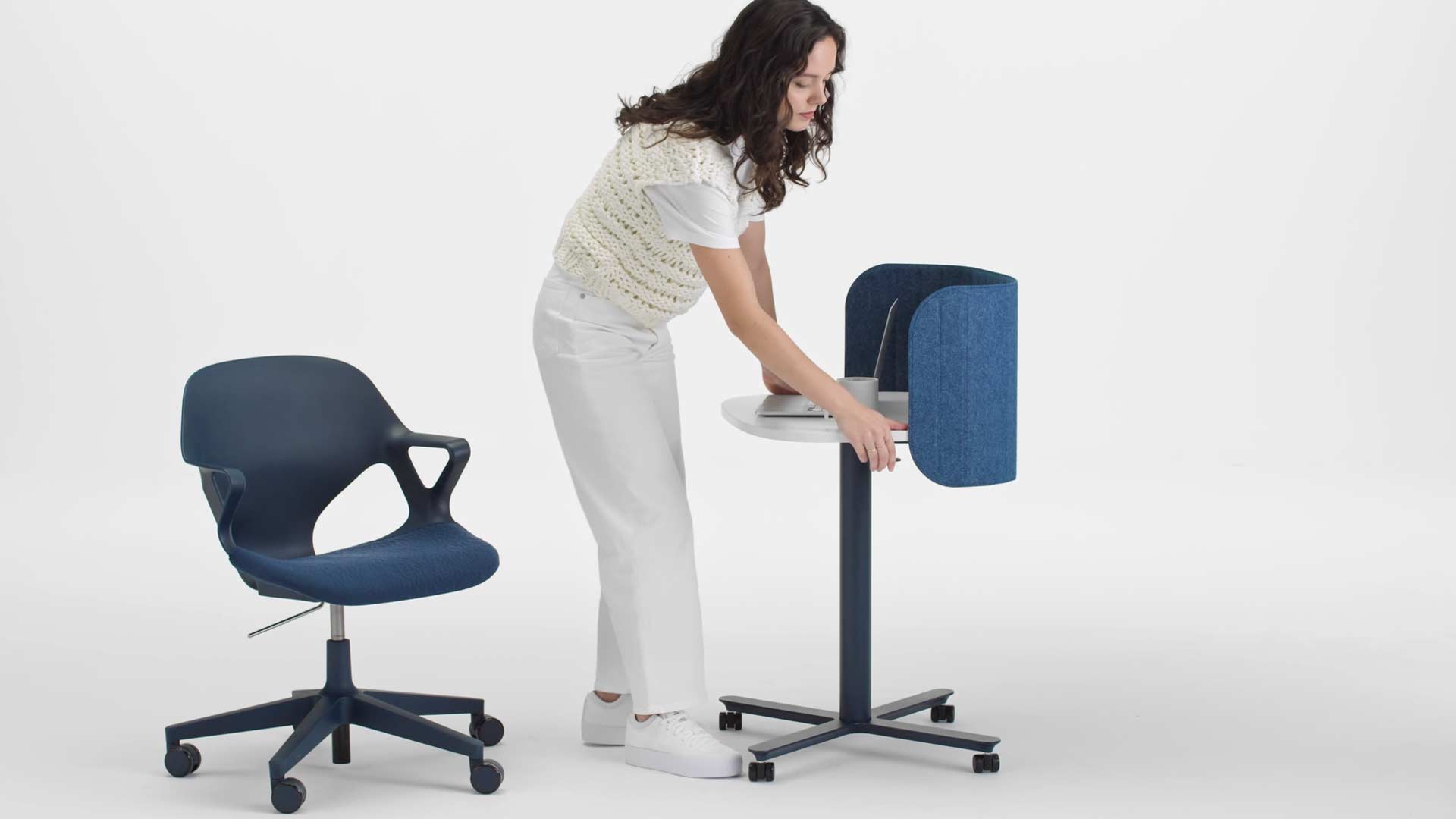 Vídeo de una mujer sentada en una silla de oficina Zeph azul levantándose y elevando la altura de una mesa de trabajo Passport azul y blanca de Herman Miller, utilizando la palanca del lateral.