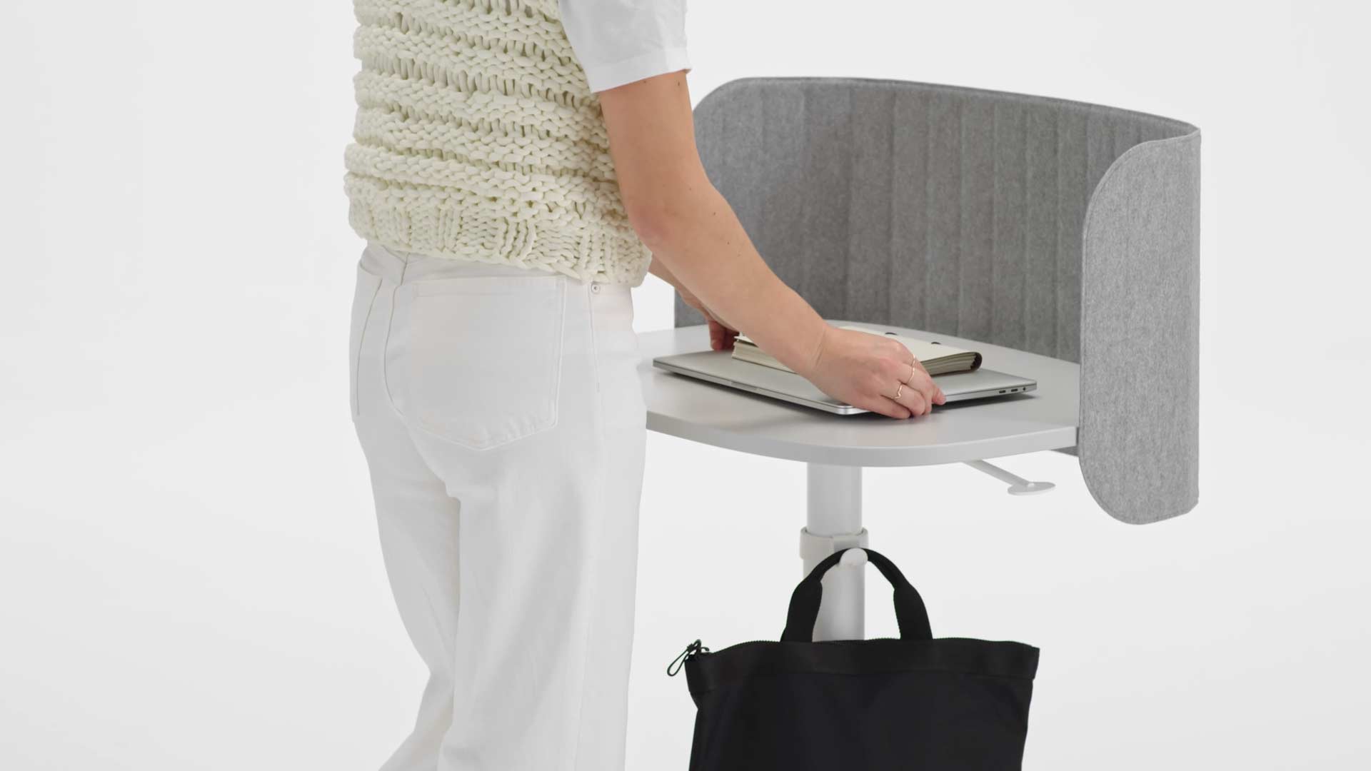 Un vídeo de una mujer utilizando una mesa de trabajo Passport blanca. Se acerca a la mesa, cuelga su bolso en el gancho para bolsos, coloca su portátil en el tablero y, a continuación, eleva la mesa a la altura correcta.