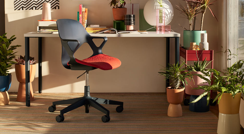 Una silla de oficina Zeph negra con un cojín de asiento rojo se coloca delante de un escritorio Herman Miller OE1. En un entorno de oficina doméstica, con un carrito OE1 rojo y una selección de plantas de interior en macetas de colores.