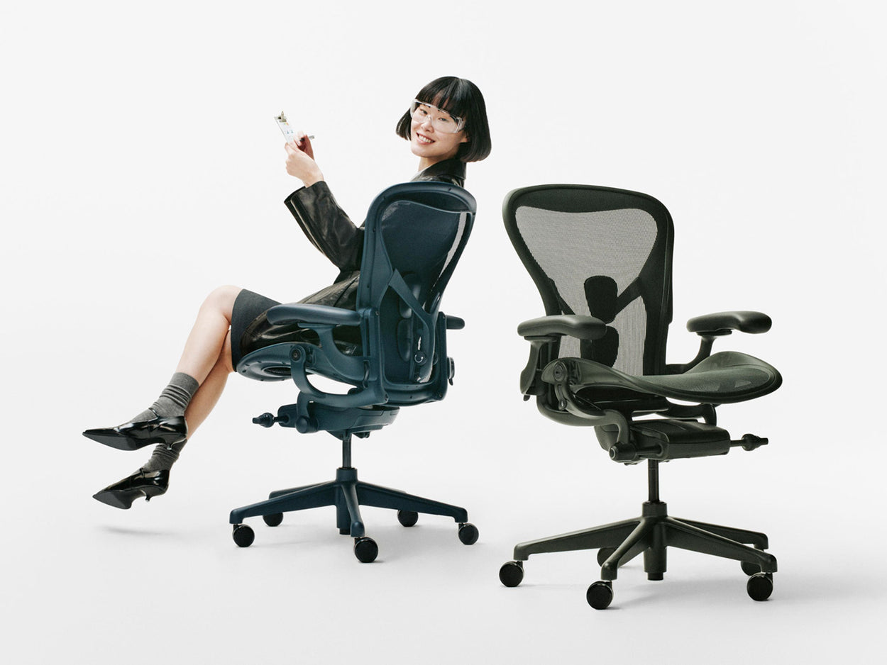Una mujer estaba sentada en una silla ergonómica de oficina Herman Miller Aeron de color azul Nighfall, junto a otra Aeron de color verde Jasper, en un estudio blanco.