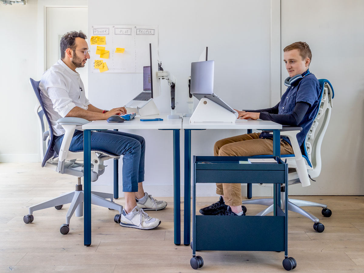 oto de dos hombres sentados en escritorios enfrentados, mostrando que cuando se está sentado correctamente, los codos quedan a la altura de la parte superior del escritorio. Características: escritorio OE1 con patas azules y tablero blanco, junto con Mirra 2 en blanco mineral y azul y Embody en blanco y azul