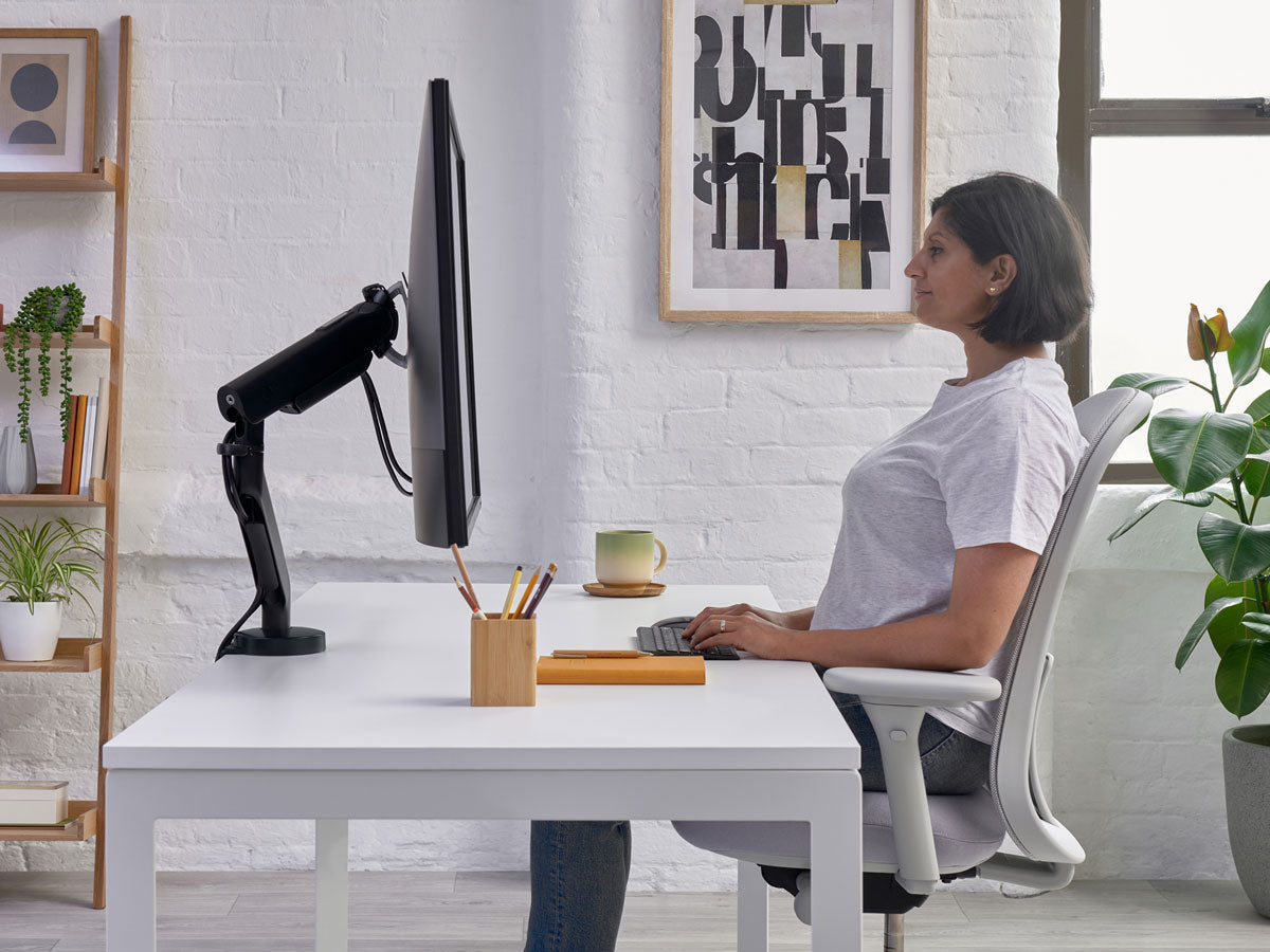 Mujer sentada en un escritorio en una silla de oficina Lino en blanco mineral con asiento gris, mostrando el nivel de las muñecas sobre el escritorio como parte de una postura correcta