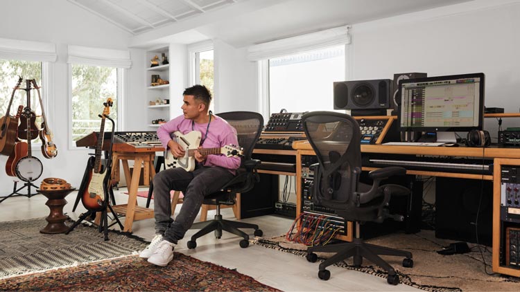 Fotografía de Rostam, músico, productor y compositor, en su estudio musical tocando la guitarra sentado en una silla de oficina Aeron en grafito