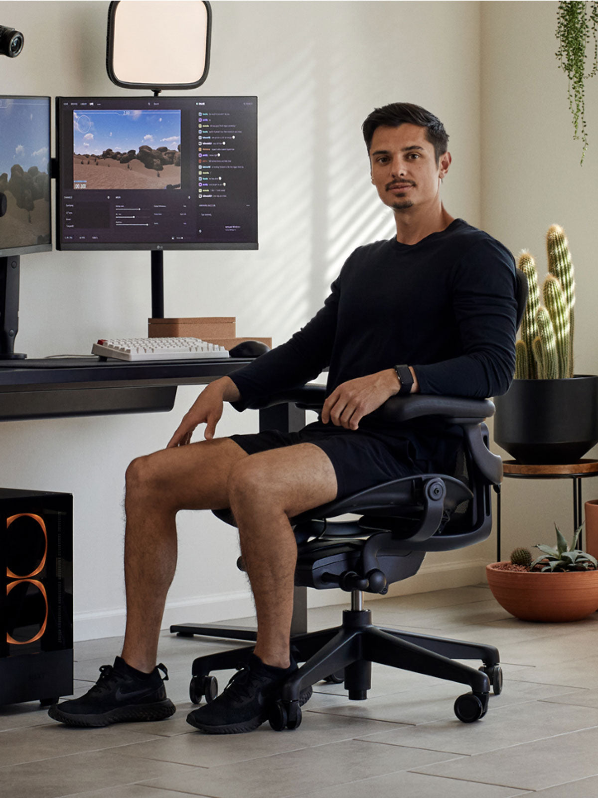 Un hombre sentado en una silla de juego Aeron negra de Herman Miller, detrás de un escritorio Nevi negro con un brazo de monitor y una pantalla Ollin negros. Ambientado en un espacio interior con suelos de baldosas, paredes blancas y plantas de interior en macetas de terracota.