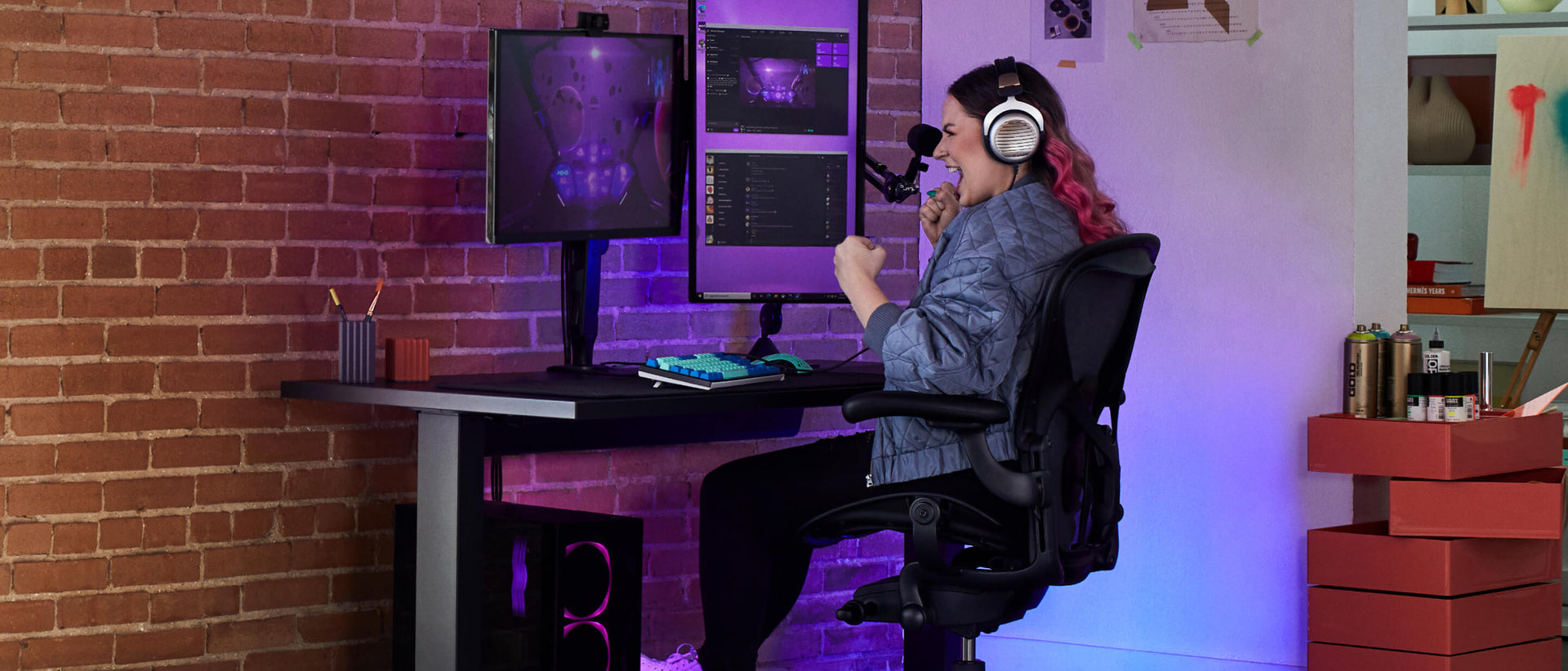 Electra sonriendo con los puños cerrados mientras juega sentada en una silla gaming Aeron en negro ónix con un escritorio gaming Nevi en negro y un brazo para monitor Ollin