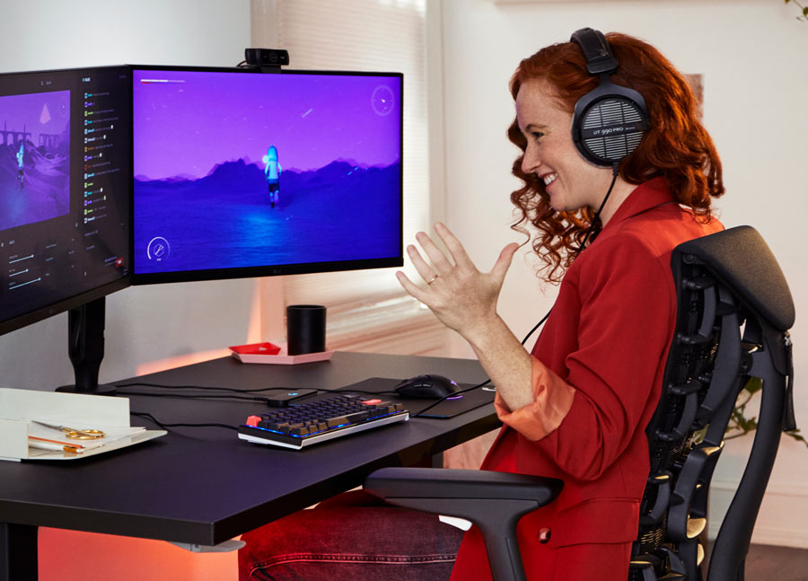 Foto de la Dra. Lindsey Migliore sonriendo mientras juega a un videojuego en la silla gaming Embody cyan con el escritorio gaming Nevi en negro.