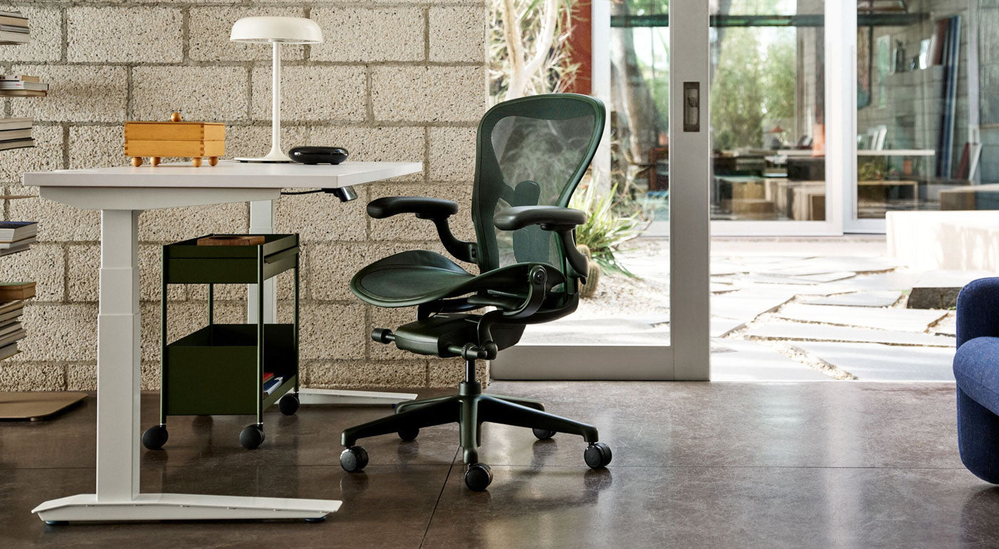 Una silla de escritorio Herman Miller Aeron en verde Jasper como parte de una luminosa oficina en casa que cuenta con un escritorio regulable en altura Jarvis blanco y un carrito de almacenamiento OE1 en verde oliva.