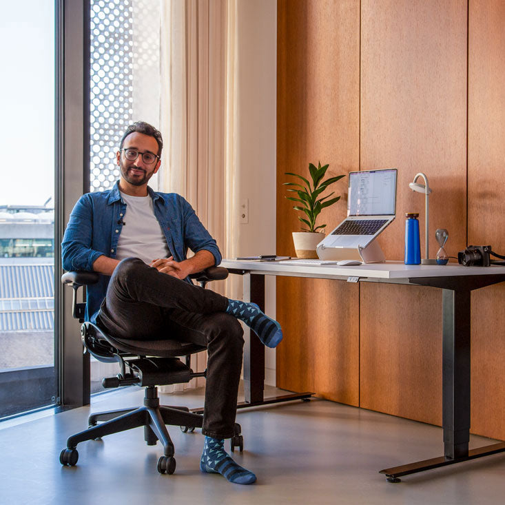 Foto de Ali Abdaal, podcaster, sentado en una silla de oficina Herman Miller Aeron en grafito delante de un escritorio Nevi sit-stand con patas negras y tapa blanca con un ordenador portátil en un soporte para portátiles Oripura