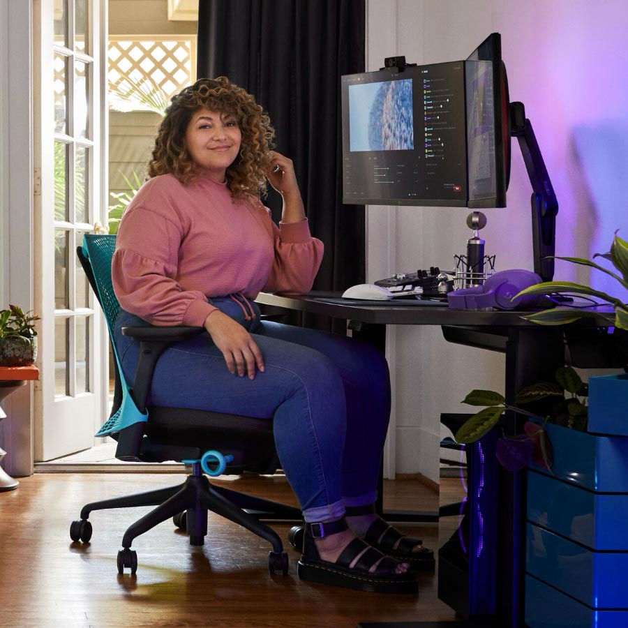 El streamer profesional y actor de doblaje Friskk sentado en la silla gaming Sayl de Herman Miller en azul océano profundo y negro, frente a un escritorio gaming Nevi negro con monitor sobre un brazo portemonitor Oliin.