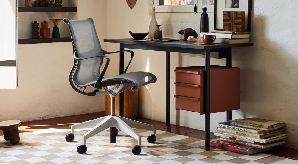 Un escritorio Mode negro de Herman Miller, con almacenamiento de terracota con manijas de nogal, se coloca frente a una silla de oficina Setu gris de Herman Miller. Un despacho bien amueblado con estanterías, libros y accesorios decorativos para el hogar.