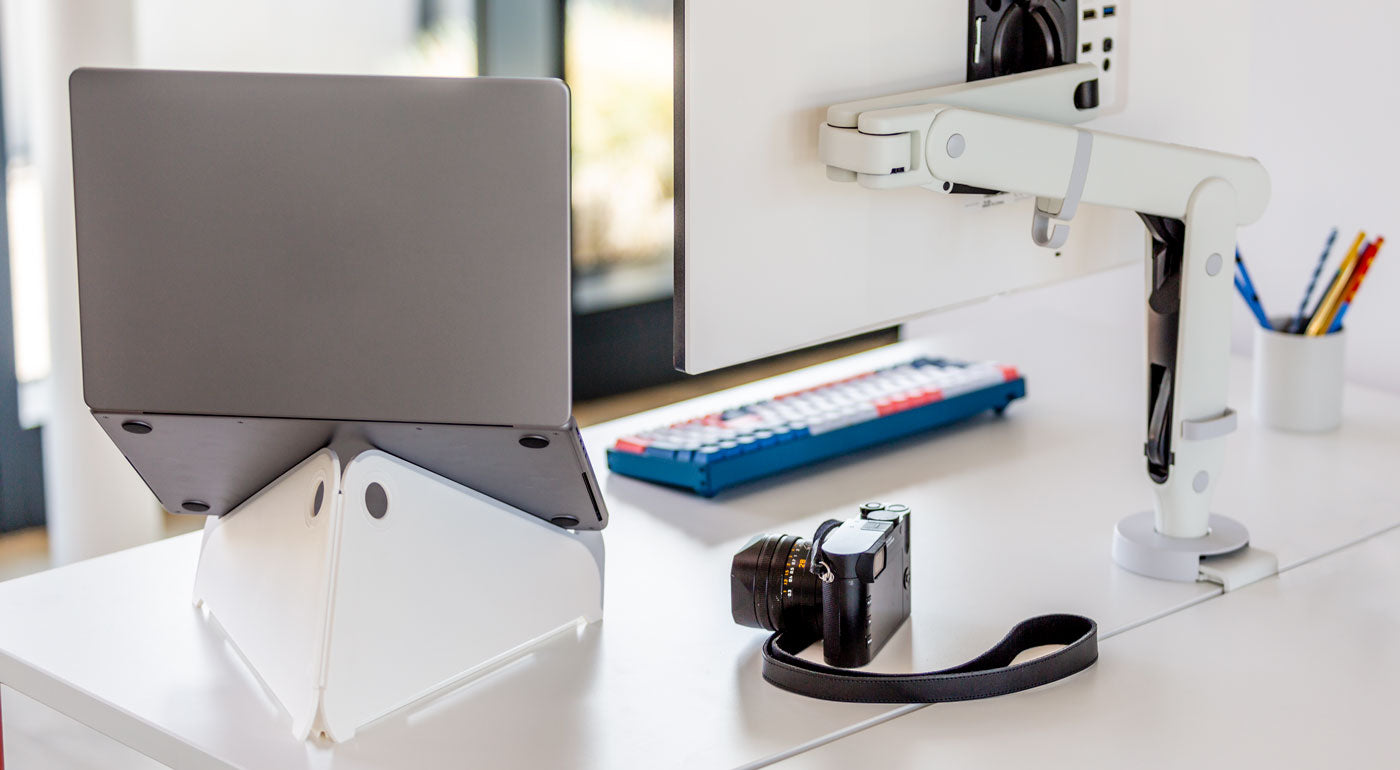 Un soporte para portátil Oripura blanco de Herman Miller sostiene un portátil junto a un brazo para monitor blanco.