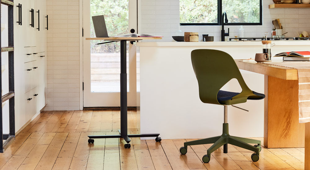 Una mesa Passport negra y de roble de Herman Miller colocada cerca de una isla de cocina blanca, con un ordenador portátil y un libro. La imagen también muestra una silla Zeph verde y negra de Herman Miller colocada en una mesa de comedor de madera.