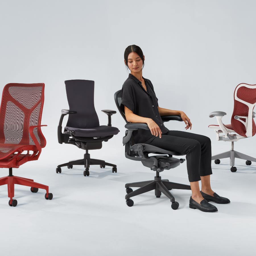Alineación de cuatro sillas de oficina Herman Miller, incluyendo Cosm con respaldo medio en rojo cañón, Embody en negro, Aeron en grafito con una mujer sentada y, por último, Mirra 2 con estructura blanca y asiento y respaldo rojos.