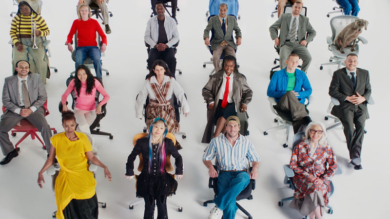 Un grupo grande y diverso de personas sentadas en una selección de sillas de oficina ergonómicas Herman Miller en un estudio blanco.