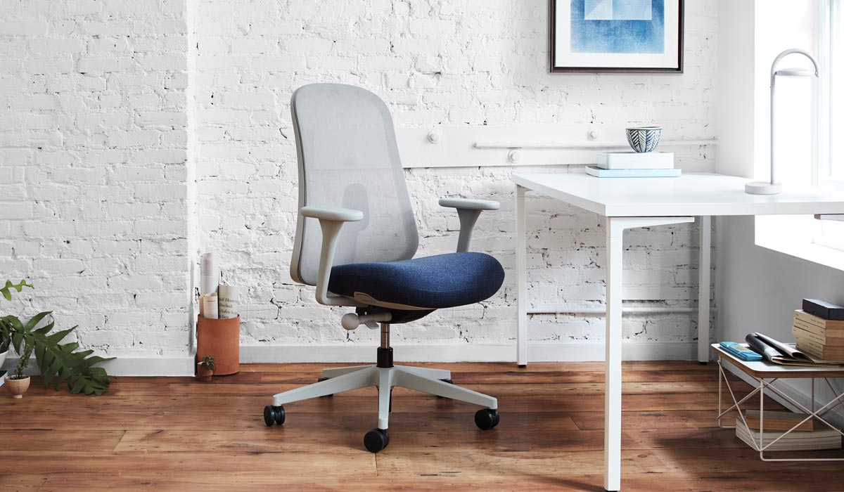 Una silla de oficina Lino gris y azul de Herman Miller, colocada delante de un escritorio blanco. El espacio interior presenta una pared de ladrillo blanco, suelo de madera y una lámpara de mesa Marselis blanca sobre el escritorio.