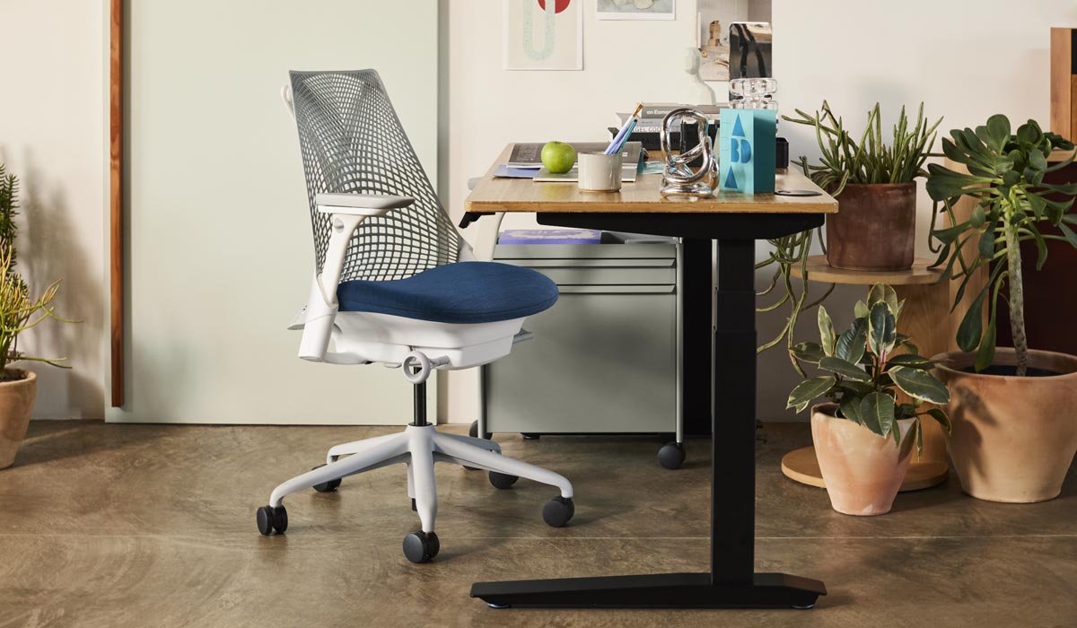 Una silla de oficina Sayl blanca y azul de Herman Miller, colocada junto a un escritorio de pie Jarvis con patas negras y encimera de bambú. En un entorno de oficina doméstica, con un carrito OE1 azul y una selección de plantas de interior en macetas de terracota.