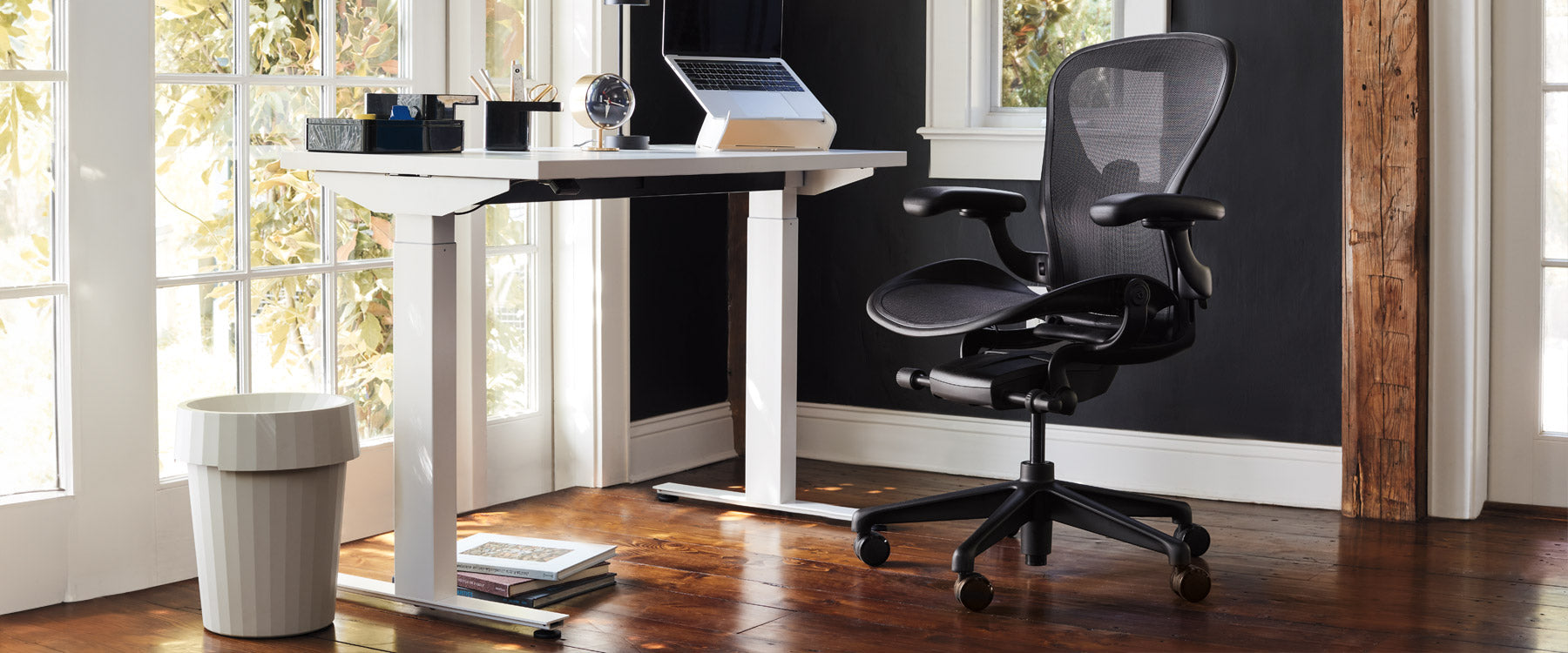 Escenario de oficina en casa con escritorio Nevi para sentarse y estar de pie en color blanco con silla de oficina Aeron en negro ónix, complementado con un soporte para portátil Oripura y una papelera Hay Shade en color blanco roto