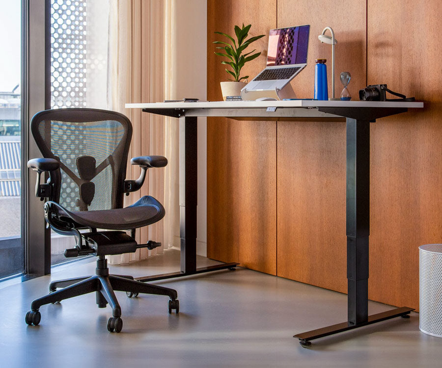 Entorno de oficina en casa que muestra el escritorio Nevi para trabajar de pie en posición vertical, con patas negras y superficie blanca, junto a una silla de oficina Aeron en color grafito al fondo.