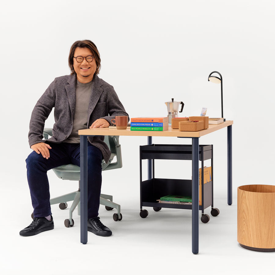 Imagen de Kevin Kwan sentado en una silla de oficina Glacier Cosm de respaldo bajo desde diferentes ángulos, delante de un escritorio OE1 de altura fija en azul noche y ceniza, con un carro de almacenamiento OE1 debajo.