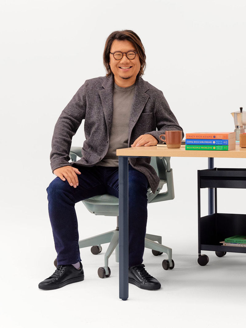 Alegre fotografía de Kevin Kwan sentado en una silla de oficina Cosm en color gris glaciar, apoyado en un escritorio OE1 de altura fija con patas en color azul noche y tablero en color ceniza claro.