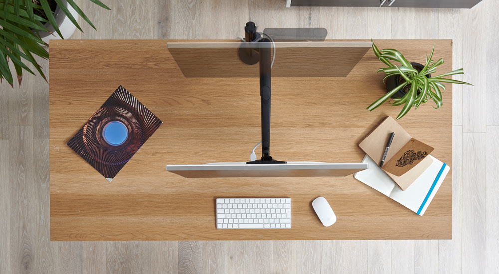 Vista superior de un brazo de monitor Lima negro de CBS fijado a un escritorio de madera, que muestra el movimiento del brazo del monitor y la pantalla.