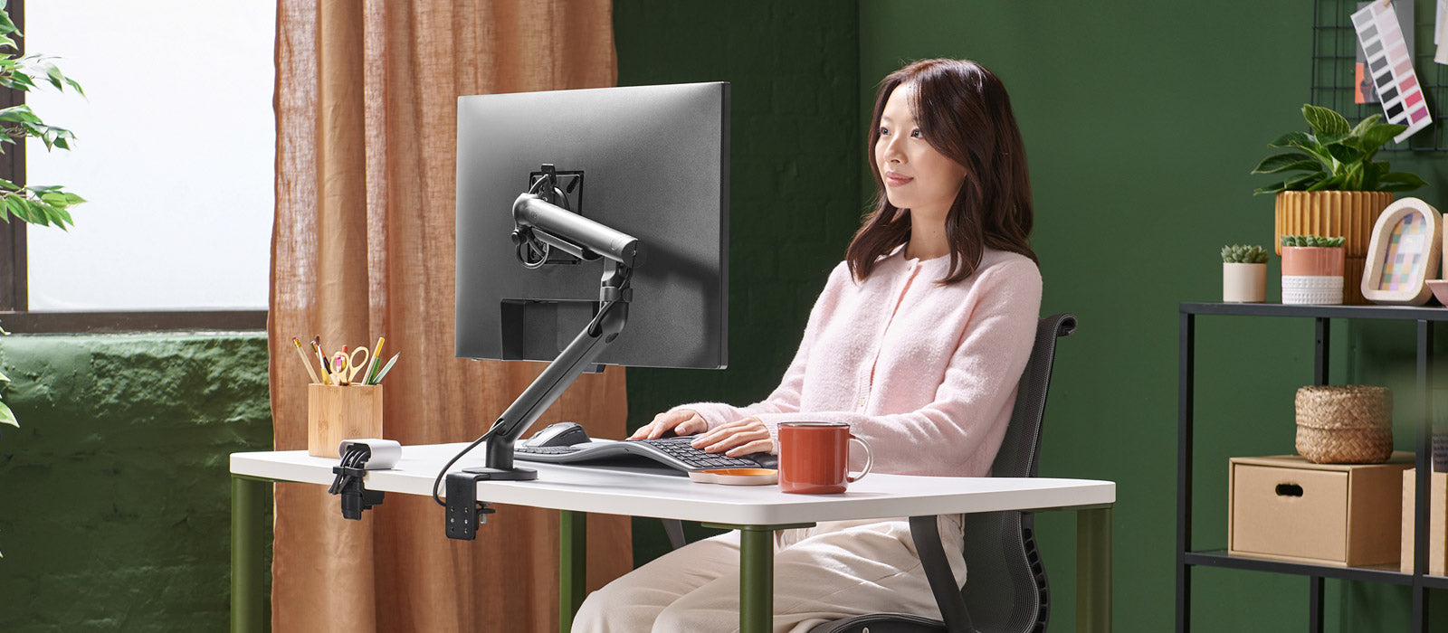 Una mujer sentada ante un escritorio Herman Miller OE1 trabajando frente a una pantalla sujeta por un brazo para monitor Flo negro.