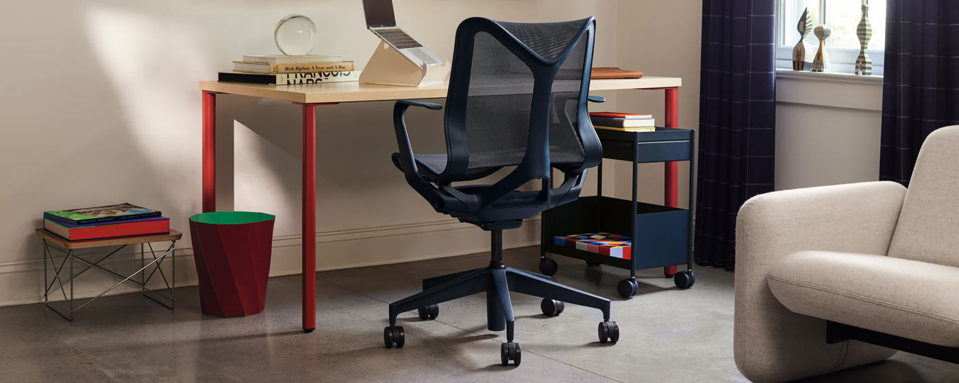 Un escritorio OE1 de madera clara con patas rojas delante de una silla de oficina ergonómica Herman Miller Cosm de color azul oscuro.