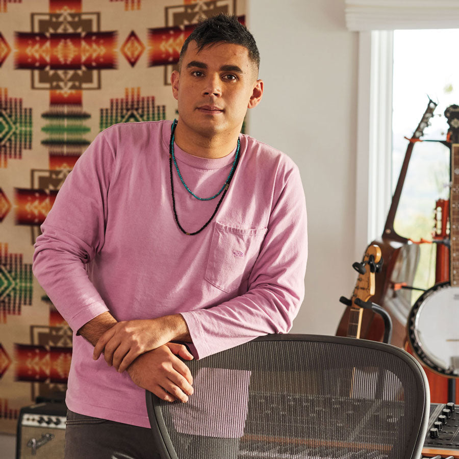 Foto de Rostam, músico, productor y compositor, de pie apoyado en una silla de oficina Aeron en grafito con instrumentos musicales al fondo