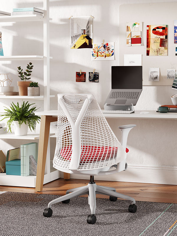 Silla de oficina para el hogar Sayl con estructura blanca, base gris y asiento tapizado en rojo en un entorno de oficina en casa, con estanterías y plantas y un ordenador portátil sobre un soporte Orpura blanco.