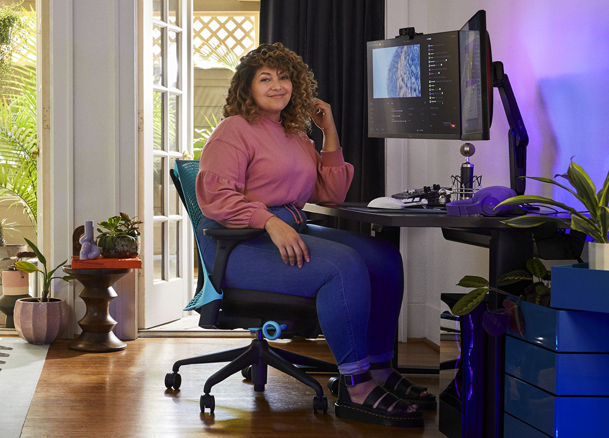 Una mujer sentada en una silla de juego Sayl azul y negra de Herman Miller, colocada detrás de un escritorio Nevi negro con un brazo de monitor y una pantalla Ollin negros. Ambientado en un espacio interior con suelos de madera, paredes blancas y plantas de interior.