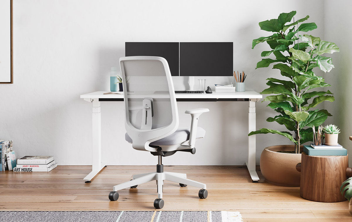 Oficina en casa con silla de oficina Verus en blanco mineral con cojín de asiento gris pálido, delante del escritorio sit-stand Nevi en blanco con dos pantallas en brazos para monitor.