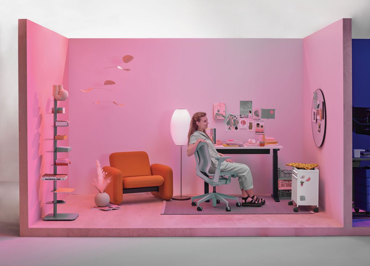 Escritorio doméstico con una mujer sentada en una silla de oficina Cosm de respaldo bajo en color gris glaciar con un escritorio compacto de pie y una silla Wilkes en el fondo.