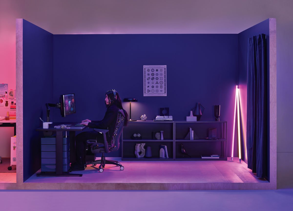 Escritorio para juegos con una mujer jugando sentada en una silla para juegos Logitech Embody en color negro con un escritorio para juegos Nevi en color negro y una lámpara de neón Hay en la esquina.