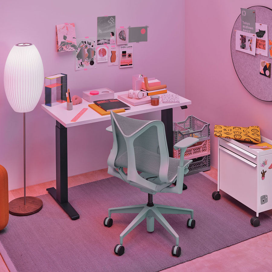 Entorno de oficina en casa con silla de oficina Cosm con respaldo bajo en color gris glaciar delante de un escritorio sit-stand Nevi con patas negras y superficie blanca.